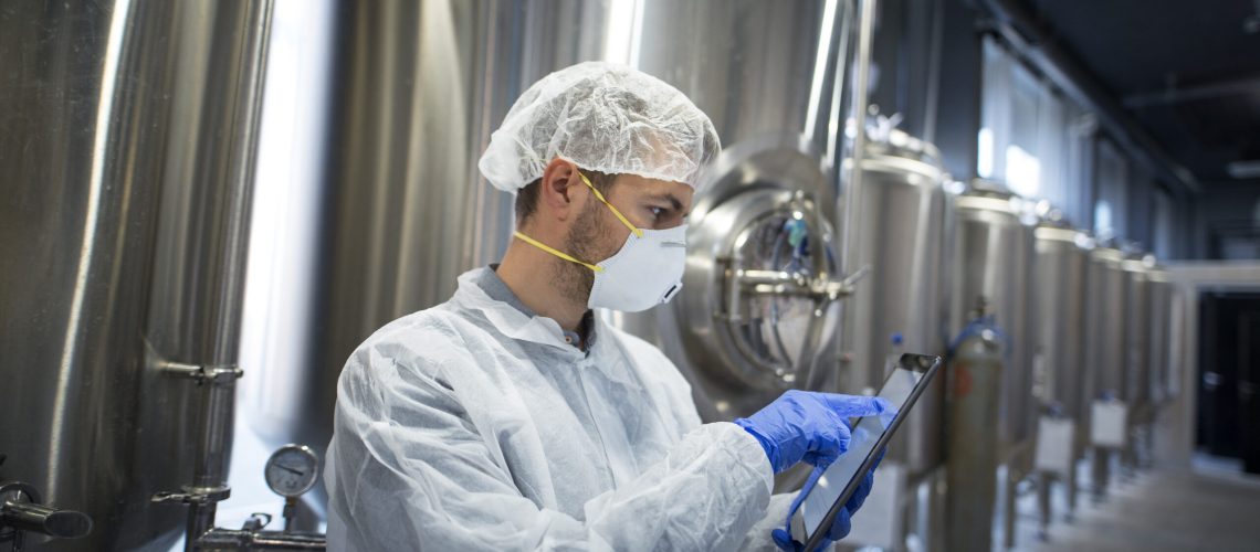 Technologist in protective suit holding tablet and checking production in factory plant.